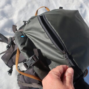 Vrchní kapsa Thule Stir Alpine je dobře přístupná pro věci rychlé potřeby. Je nepromokavá. Oranžová poutka dobře dlouhá k snadné manipulaci s batohem.